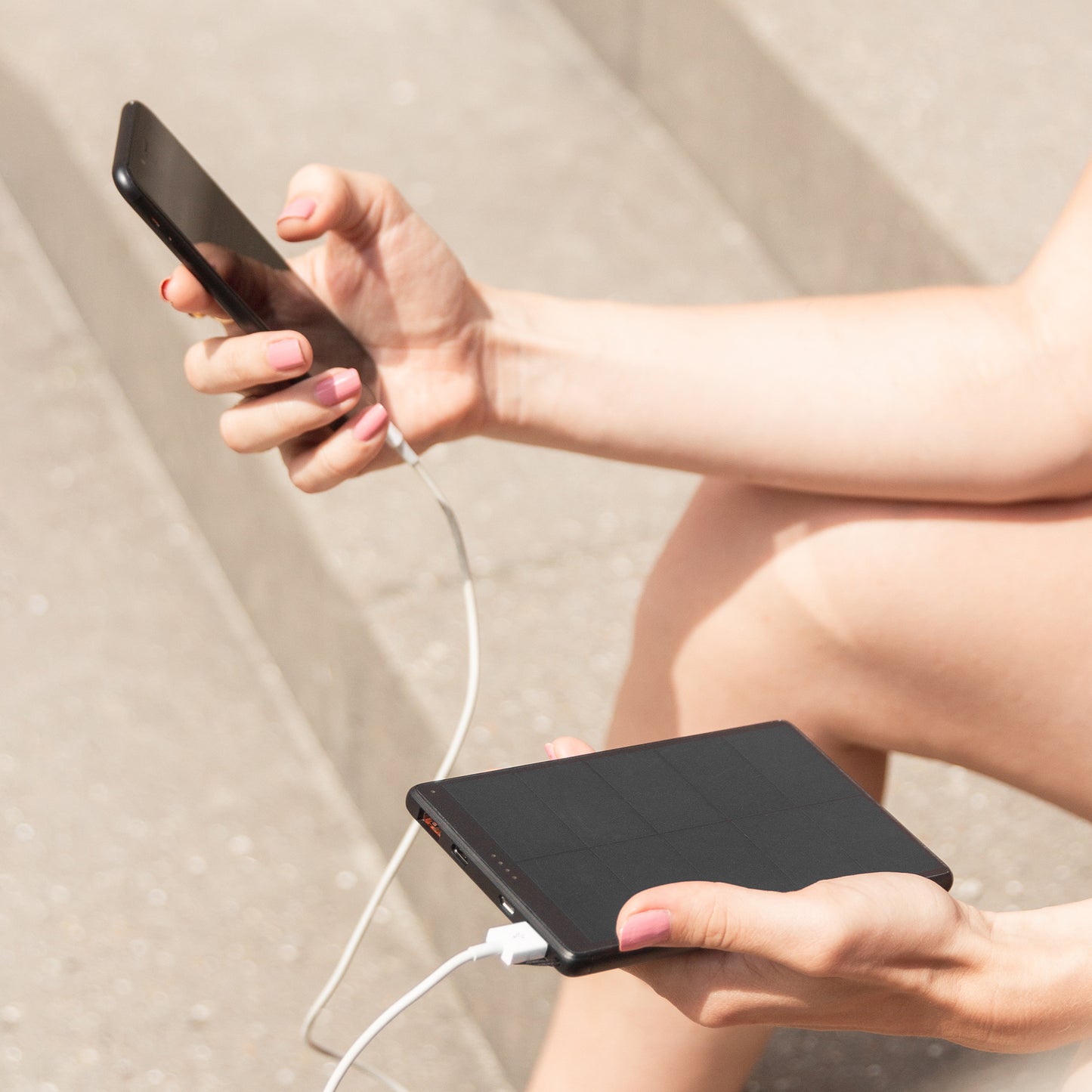 Frau Sitzt auf Treppen, hält Sandberg Urban Solar Powerbank 10000 ohne Verpackung in der Hand und lädt ihr iPhone
